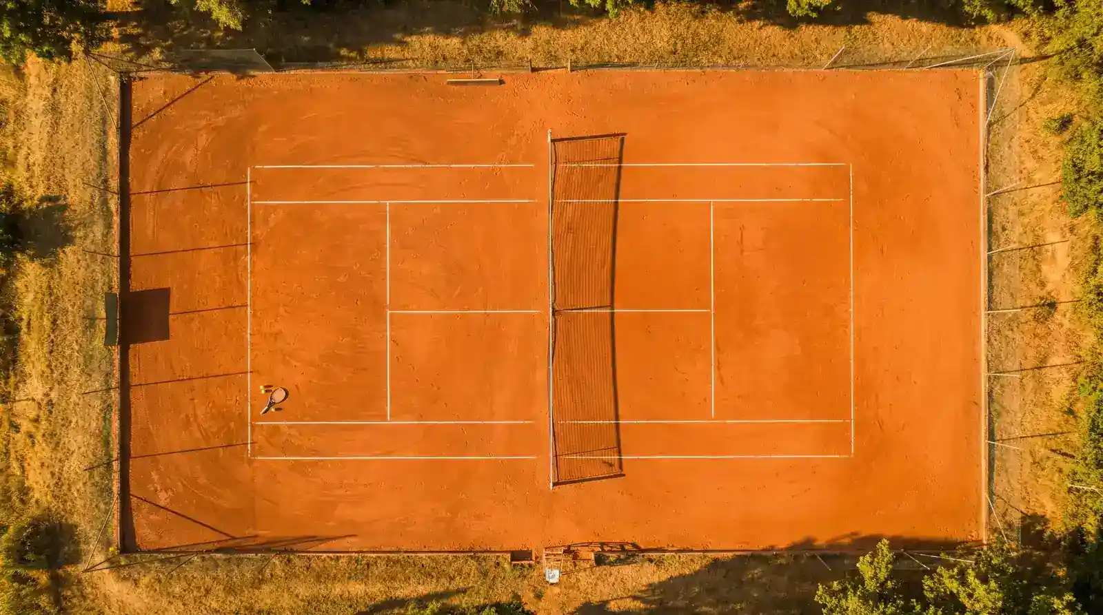 Pista de tenis de tierra batida vista desde arriba con líneas marcadas y sombras alargadas